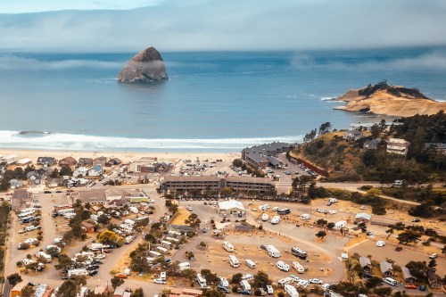 A coastal view featuring a rocky outcrop, beach, and a parking area with RVs near a small town.