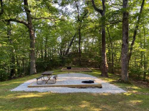 A serene outdoor space with a picnic table, grill, and gravel area surrounded by lush green trees.