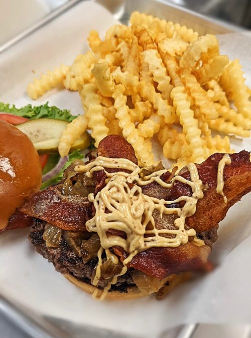 A juicy burger topped with bacon and sauce, served with crinkle-cut fries and a side of lettuce and tomato.