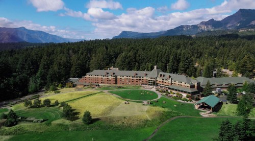 Aerial view of a large lodge surrounded by lush green fields and mountains under a partly cloudy sky.