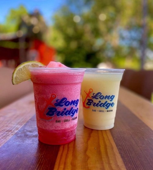 Two colorful frozen drinks in cups with "Long Bridge Bar & Grill" logo, sitting on a wooden table outdoors.