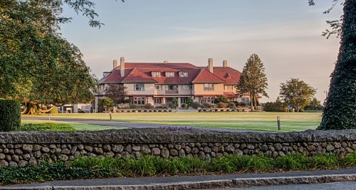 A large, elegant mansion with a red roof, surrounded by lush greenery and a stone wall in the foreground.