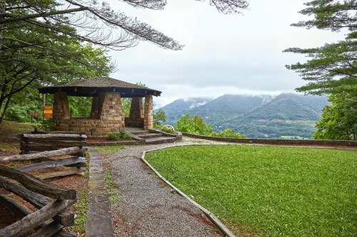 A stone gazebo surrounded by greenery, with mountains and cloudy skies in the background.