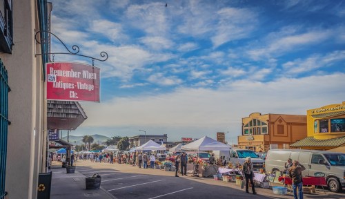 A bustling outdoor market with tents, vendors, and shoppers under a blue sky, featuring a sign for an antique shop.
