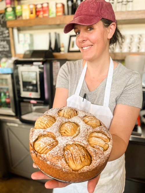 A smiling woman in a cap holds a freshly baked apple cake dusted with powdered sugar in a cozy café setting.