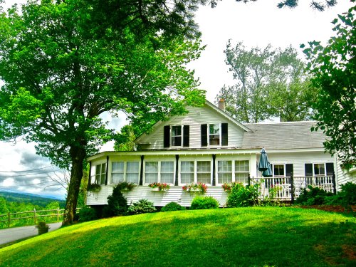 A charming white house with a porch, surrounded by lush greenery and trees under a partly cloudy sky.