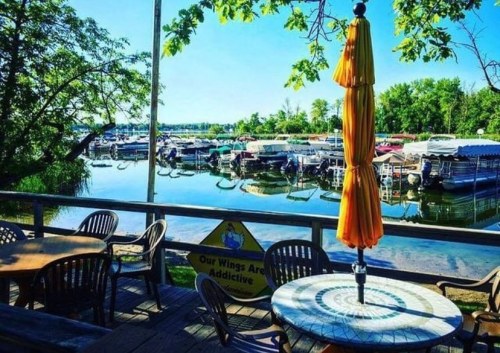 A scenic view of a marina with boats, surrounded by trees and a sunny blue sky, seen from a wooden deck with a table.