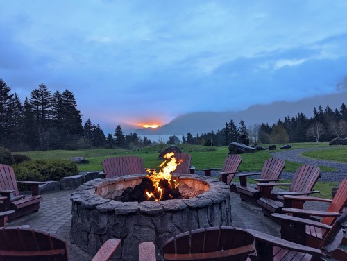 A cozy fire pit surrounded by wooden chairs, with a scenic view of trees and mountains at sunset.