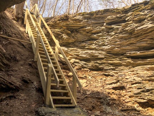 A wooden staircase leads up a rocky cliff in a wooded area, surrounded by trees and natural stone formations.