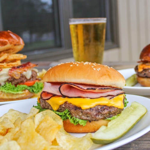 A close-up of a cheeseburger with ham, lettuce, and pickles, served with fries and a glass of beer in the background.