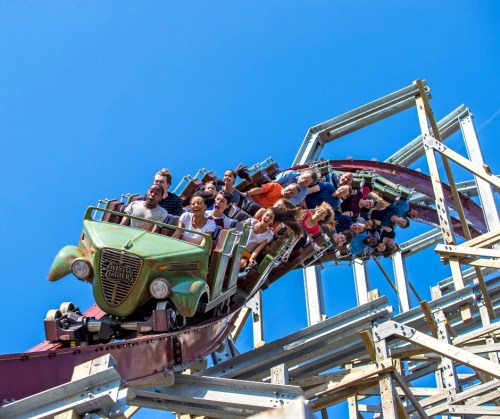 A roller coaster with a vintage car design twists through the air, filled with excited riders against a clear blue sky.