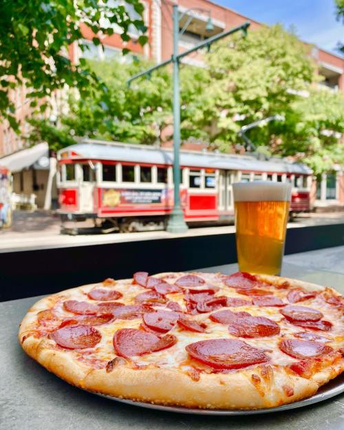 A pepperoni pizza on a table with a glass of beer, set against a backdrop of a red trolley and green trees.