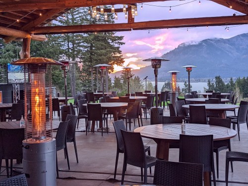 Outdoor dining area with tables and heaters, overlooking a lake and mountains at sunset.