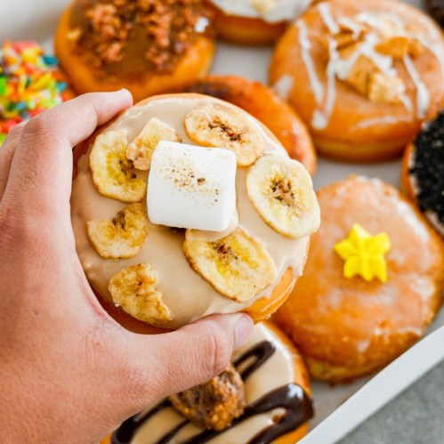 A hand holding a donut topped with banana chips and a marshmallow, surrounded by various colorful donuts.