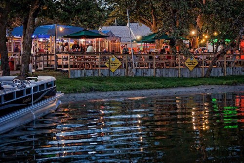 A waterfront dining area with colorful lights, tables, and a boat docked nearby, reflecting in the water at dusk.
