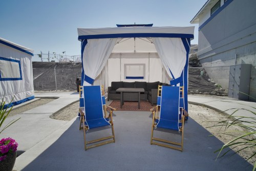 A cozy outdoor seating area with a tent, two lounge chairs, and a small table, surrounded by gravel and plants.