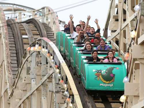 A group of excited people on a roller coaster, hands raised, as they descend a steep track at an amusement park.