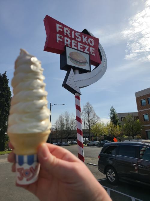 A hand holds a soft-serve ice cream cone in front of the Frisko Freeze sign on a sunny day.