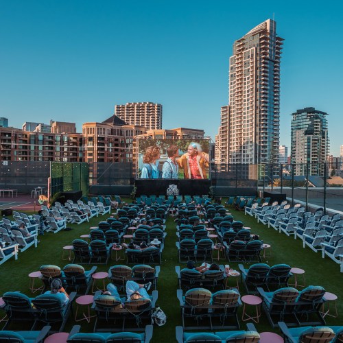 Outdoor movie screening with lounge chairs set up on grass, large screen displaying a film, and city skyline in the background.