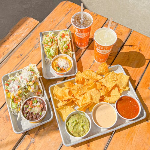 A vibrant spread of tacos, salads, chips, guacamole, and drinks on a wooden table.