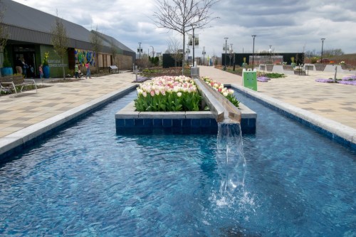 A serene water feature flows through a landscaped area with blooming tulips and modern seating under a cloudy sky.