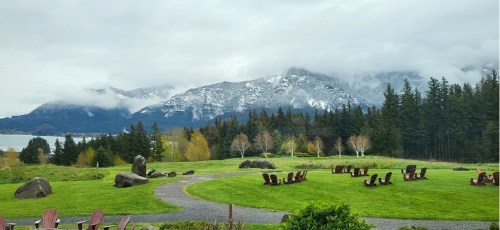 A scenic view of mountains with snow, surrounded by green grass and trees, featuring a path and wooden chairs in the foreground.