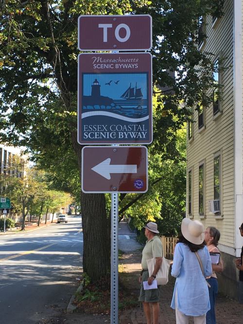 Sign for Essex Coastal Scenic Byway with an arrow pointing left, surrounded by trees and people walking nearby.