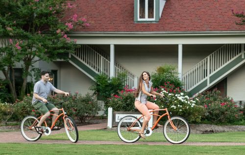 A man and woman ride orange bicycles on a grassy area near a house with blooming flowers and stairs in the background.