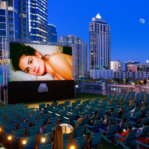 Outdoor movie screening with a large screen showing a film, surrounded by modern buildings and a clear night sky.