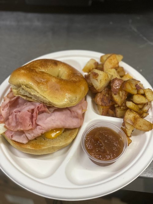 A plate with a bagel sandwich filled with ham and cheese, alongside seasoned potato cubes and a small cup of salsa.