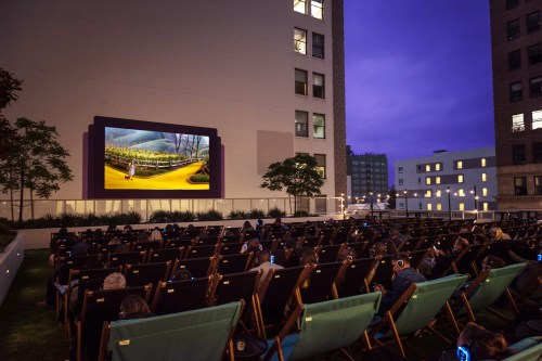 Outdoor movie screening at dusk, with people seated in deck chairs, watching a film on a large screen.