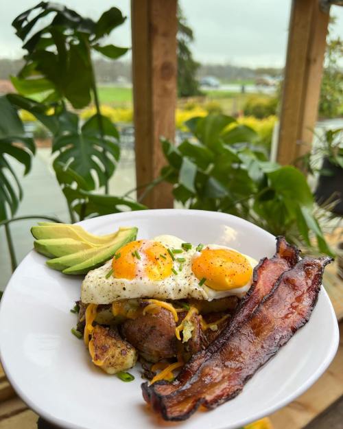 A plate of breakfast featuring two eggs, bacon, avocado, and seasoned potatoes, surrounded by greenery.