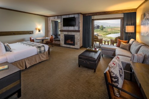 Cozy hotel room with a king bed, seating area, fireplace, and large windows overlooking a scenic landscape.