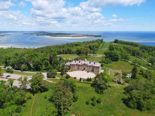 Aerial view of a large mansion surrounded by lush greenery and a scenic waterfront landscape under a blue sky.