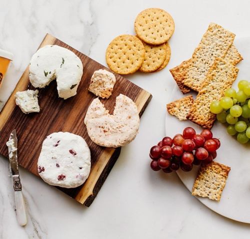 A wooden board with various cheeses, crackers, grapes, and a knife on a marble surface.