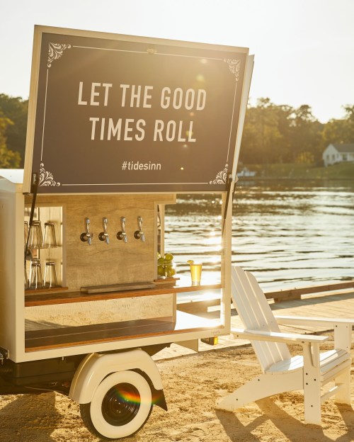 A mobile bar with a sign reading "Let the Good Times Roll" by a serene lakeside, with a sandy area and chairs.
