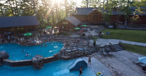 Aerial view of a pool area with people swimming, surrounded by trees and a lodge in the background, under a sunset.