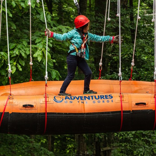 A person in a helmet stands on a raft suspended by ropes in a forested area, ready for an adventure.