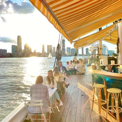 A vibrant sunset view from a boat with people dining and a city skyline in the background.