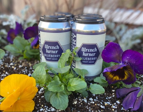 Two cans of Wrench Turner Blueberry Draft Mead nestled among colorful flowers in a garden setting.