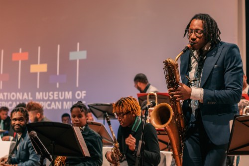 A musician plays saxophone on stage at the National Museum of African American Music, with an ensemble in the background.