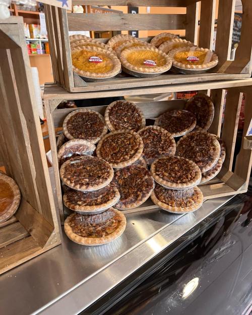 A display of various pies stacked in wooden crates, featuring pecan and other flavors, in a cozy shop setting.