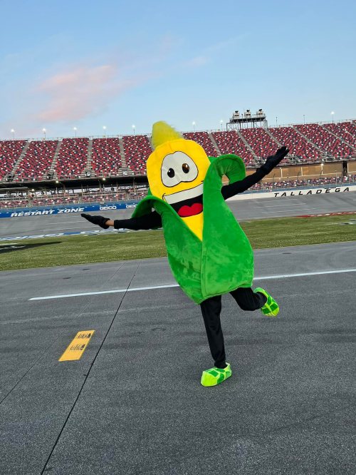 A person in a corn mascot costume joyfully poses on a racetrack with a stadium in the background.