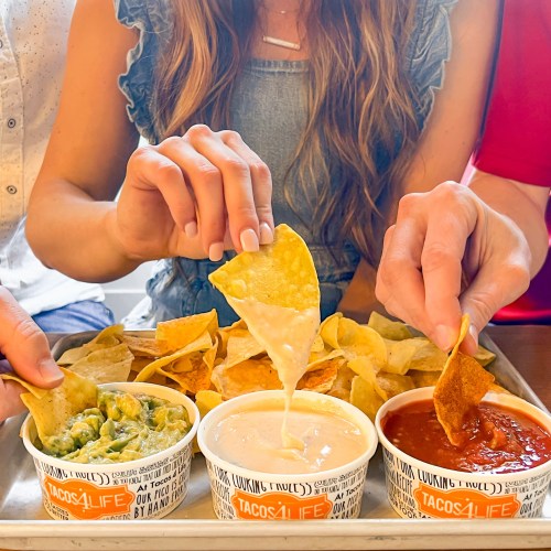 Three hands dipping tortilla chips into various sauces, surrounded by a pile of chips on a tray.
