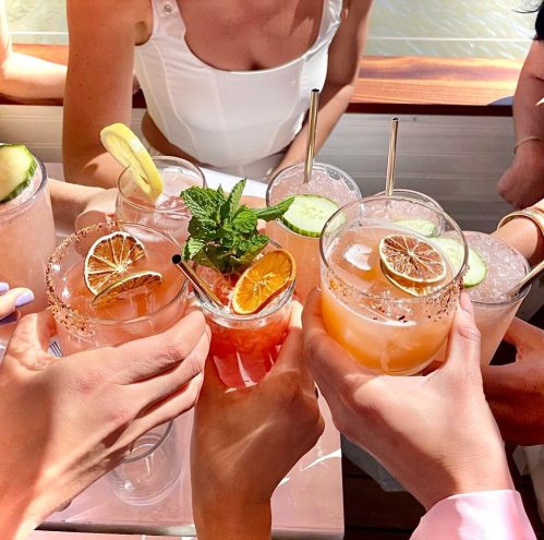 A group of hands holding colorful cocktails with garnishes, celebrating together in a sunny outdoor setting.