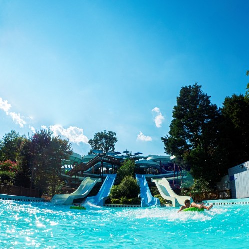 A sunny water park scene featuring two water slides and people enjoying the pool area surrounded by trees.