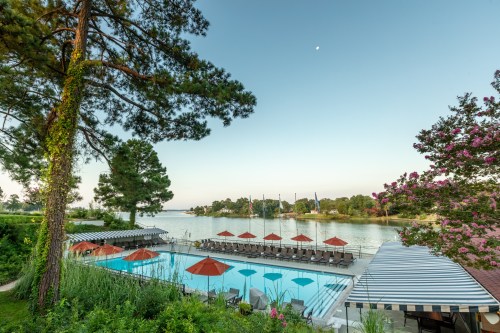 A serene lakeside view featuring a pool with orange umbrellas, surrounded by lush greenery and calm waters.