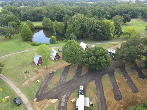 Aerial view of a campsite with red cabins, a pond, and tree-lined areas, surrounded by dirt paths and vehicles.