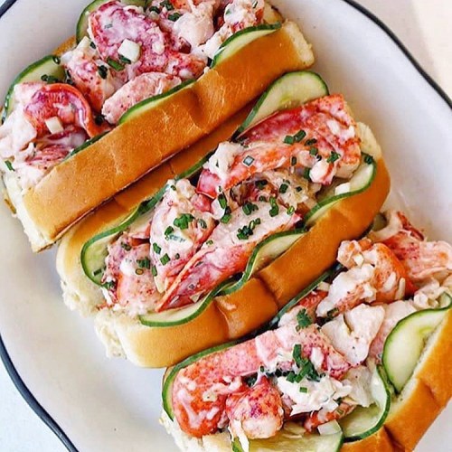 Three lobster rolls topped with cucumber slices and chives, served on a white plate.