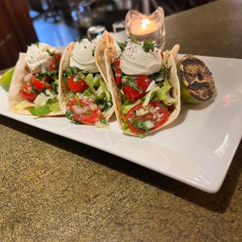 Three tacos filled with lettuce, tomatoes, and topped with sour cream, served on a white plate with a grilled lime.